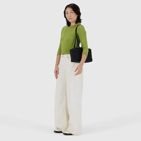 Woman wearing olive green long-sleeve top and wide-leg white pants carrying a black shoulder bag on gray background