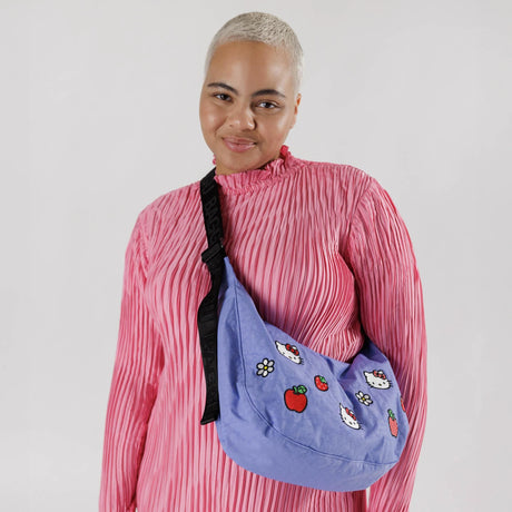 A person in a pink pleated top holds a Baggu Hello Kitty x Medium Nylon Crescent Bag, featuring whimsical Hello Kitty patterns, standing against a plain background.