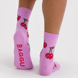Pair of light purple ankle socks with red cherries and green stems, red text on sole, worn on feet against white background