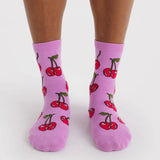 Pair of mid-calf purple socks featuring red cherries with green stems worn on feet against a white background