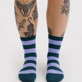 Pair of crew socks with alternating thick navy blue and lavender horizontal stripes worn on feet against white background