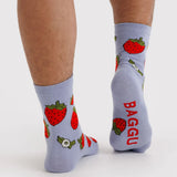 Light purple crew socks featuring red strawberries with green leaves and red text on sole worn on feet against white background