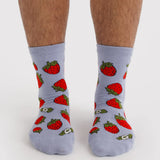 Pair of light gray crew socks adorned with bright red strawberries with green leaves on feet against a white background