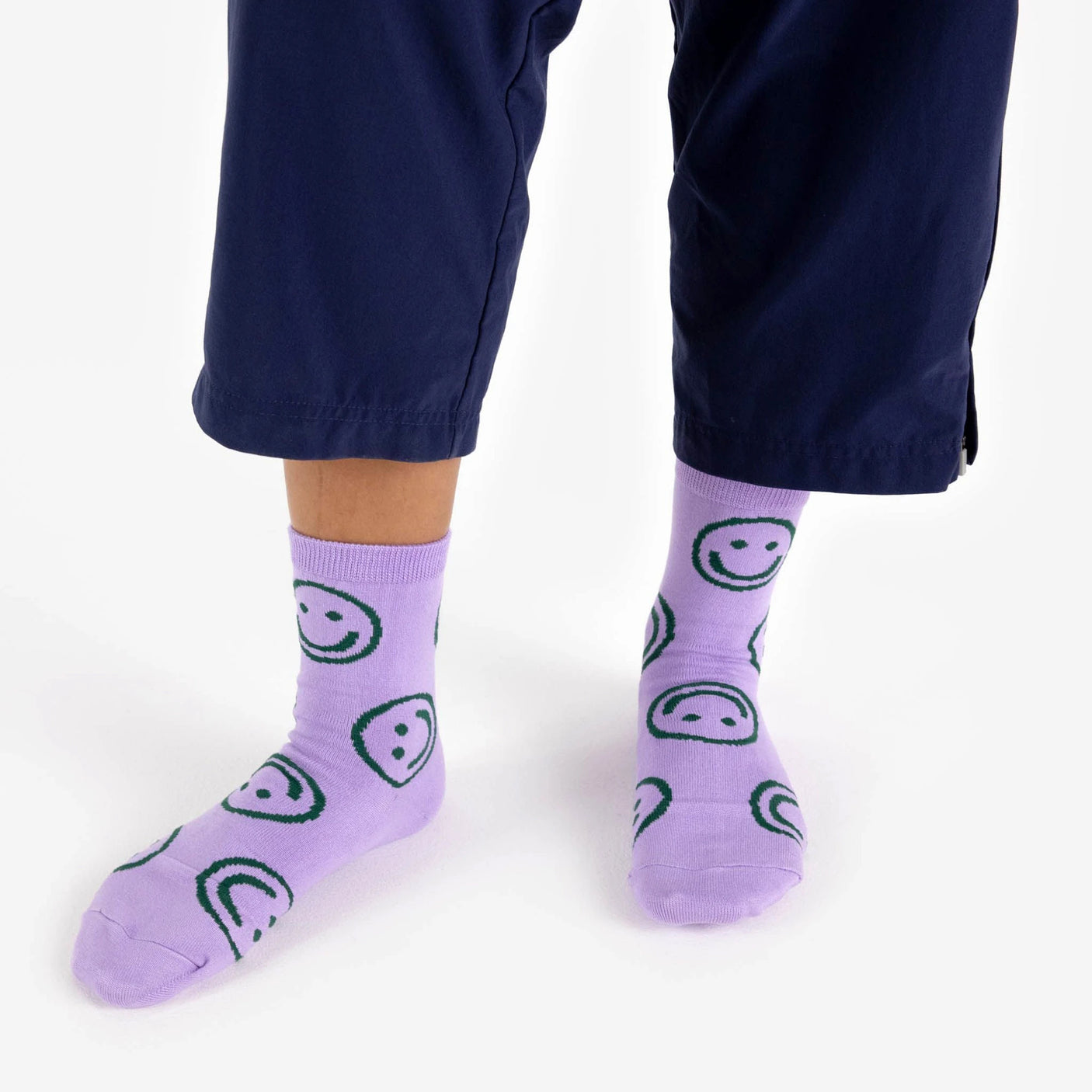 A person wearing navy pants complemented by Baggu Crew Sock - Lavender Happy, featuring playful green smiley and sad face patterns.