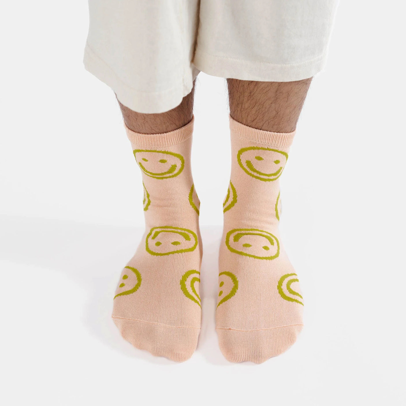 A person stands on a white background, their feet adorned with the cosy and soft Baggu Crew Sock in light pink, accented by cheerful yellow smiley faces.