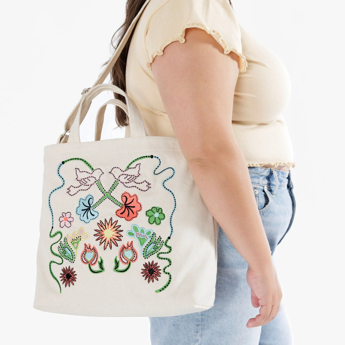 A person wearing a beige top and jeans carries a Baggu Duck Bag Medium - Embroidered Birds, showcasing vibrant floral patterns and embroidered bird designs.