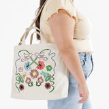 A person wearing a beige top and jeans carries a Baggu Duck Bag Medium - Embroidered Birds, showcasing vibrant floral patterns and embroidered bird designs.