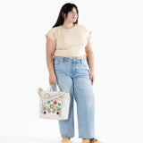 A person in a light beige top and blue jeans holds a Baggu Duck Bag Medium - Embroidered Birds, showcasing its versatility.