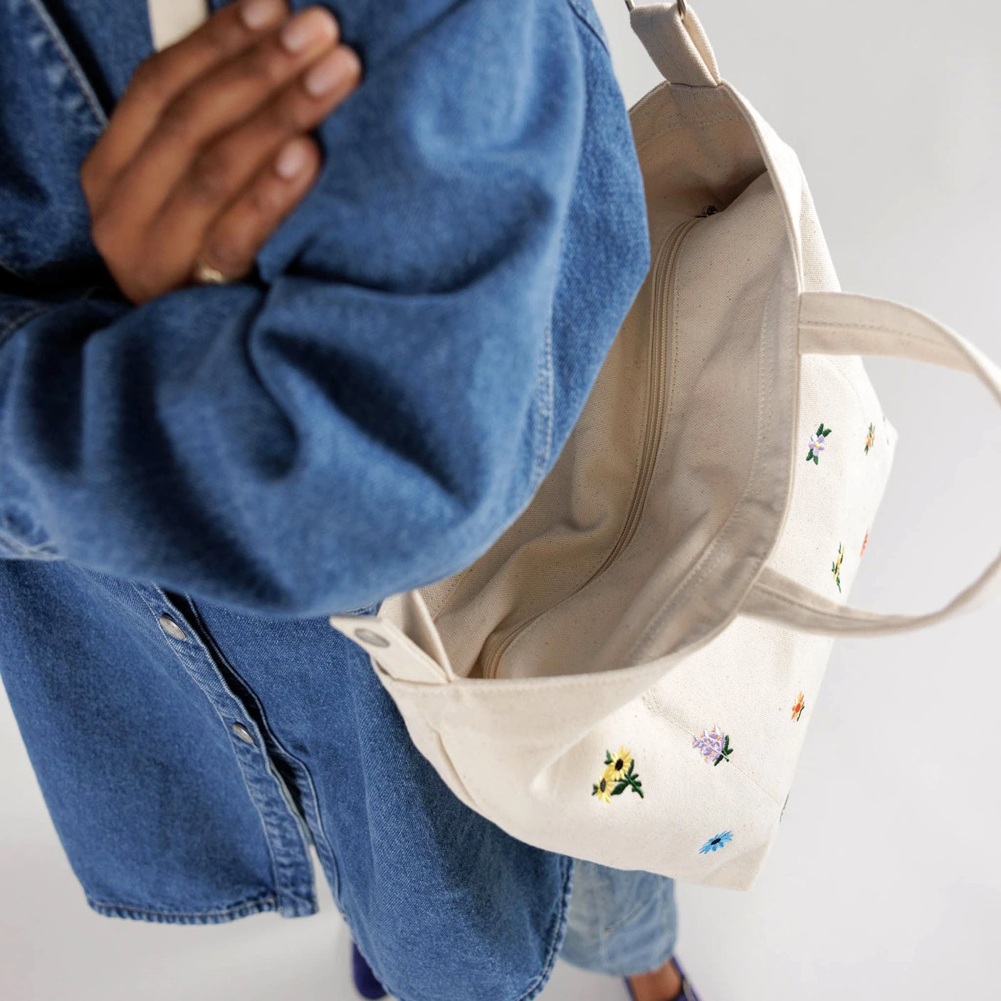 A person in a blue denim shirt is holding the Baggu Duck Bag Medium - Ditsy Floral, a beige tote bag from Baggu featuring floral embroidery and an adjustable strap.