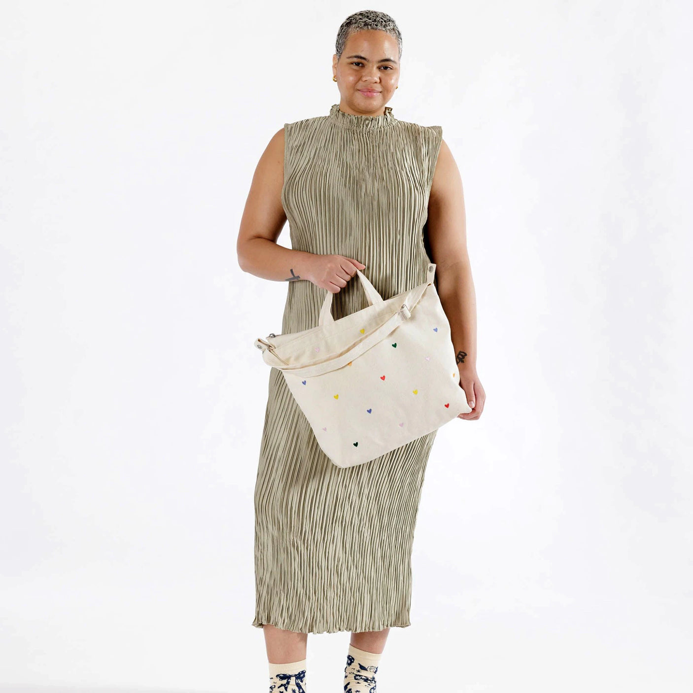 A person in a pleated dress holds the Baggu Duck Bag Medium from Baggu, which is beige and adorned with embroidered hearts and small colorful dots.