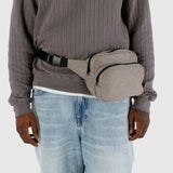 A person wearing a gray sweater and blue jeans completes their outfit with a Baggu Fanny Pack Dove, which features an adjustable strap made from durable nylon.