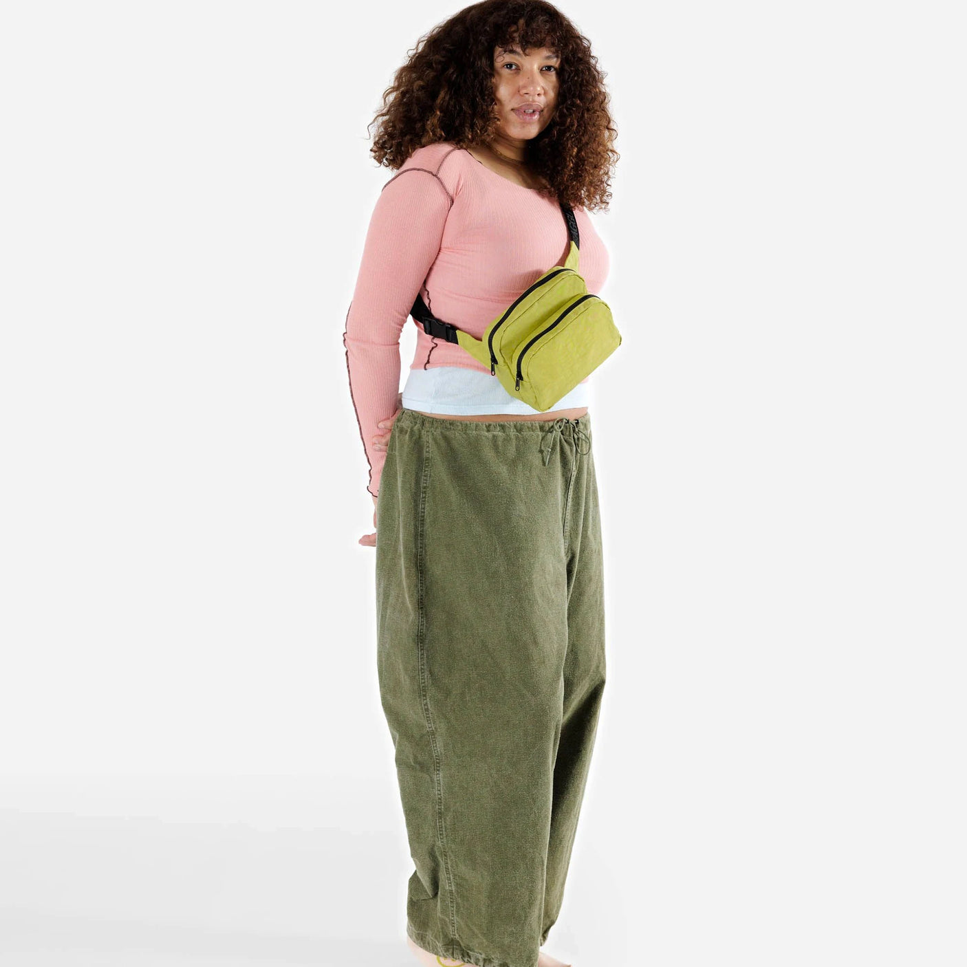 A person with curly hair is wearing a pink long-sleeve shirt, oversized green pants, and a Baggu Fanny Pack in lemongrass color. They are standing against a plain white background.