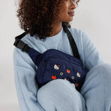 A person in a blue outfit sits cross-legged, wearing the limited edition Hello Kitty x Baggu Fanny Pack adorned with embroidered cartoon and fruit designs.