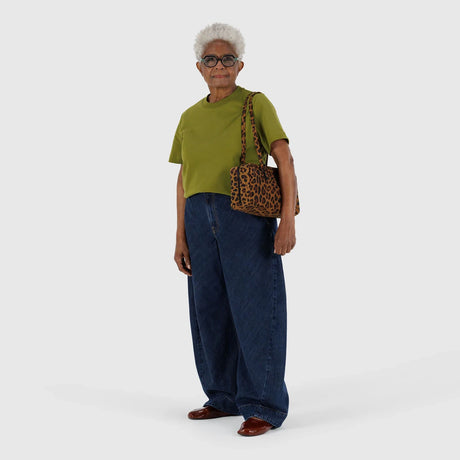 Elderly woman wearing olive green short-sleeve top, loose dark blue jeans, brown shoes, and carrying leopard print shoulder bag