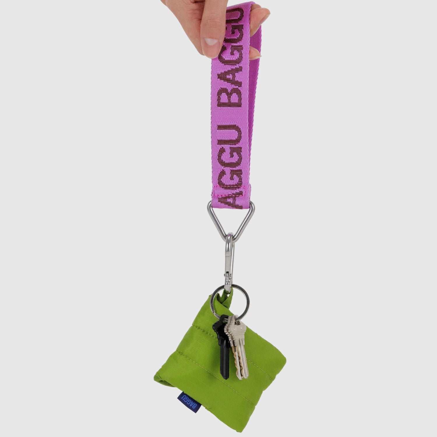 A hand holds a pink Baggu Logo Keychain with a metal clip attached to a green pouch and two keys.