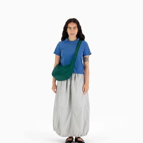 A person stands against a plain background, wearing a blue shirt and gray skirt, while carrying the Baggu Medium Nylon Crescent Bag in Cypress, featuring an adjustable tonal logo strap.