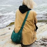 A person in a beige coat and black hoodie sits on the rocky shore with a Baggu Medium Nylon Crescent Bag in Cypress, featuring an adjustable tonal logo strap, resting beside them as they face the ocean.