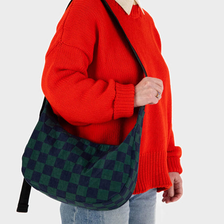 A person wearing a red sweater exudes retro-cool vibes, casually holding a Baggu Medium Nylon Crescent in Navy Green Check over the shoulder, with light blue jeans peeking through.