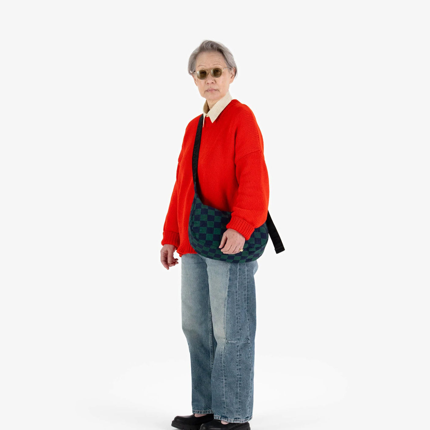 A person with short gray hair exudes retro-cool vibes, wearing sunglasses, a red sweater, blue jeans, and black shoes. They hold a Baggu Medium Nylon Crescent - Navy Green Check bag against a plain white background.