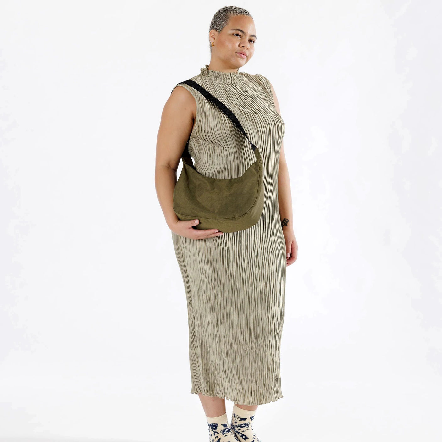 A person poses wearing a sleeveless, striped dress, featuring the Baggu Medium Nylon Crescent Bag in Seaweed, elegantly styled as a crossbody. They have a neutral expression and stand against a light background.