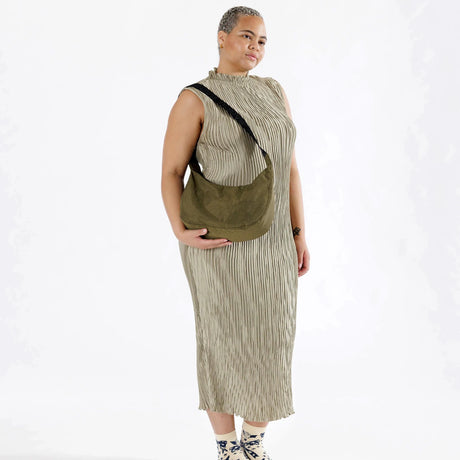 A person poses wearing a sleeveless, striped dress, featuring the Baggu Medium Nylon Crescent Bag in Seaweed, elegantly styled as a crossbody. They have a neutral expression and stand against a light background.