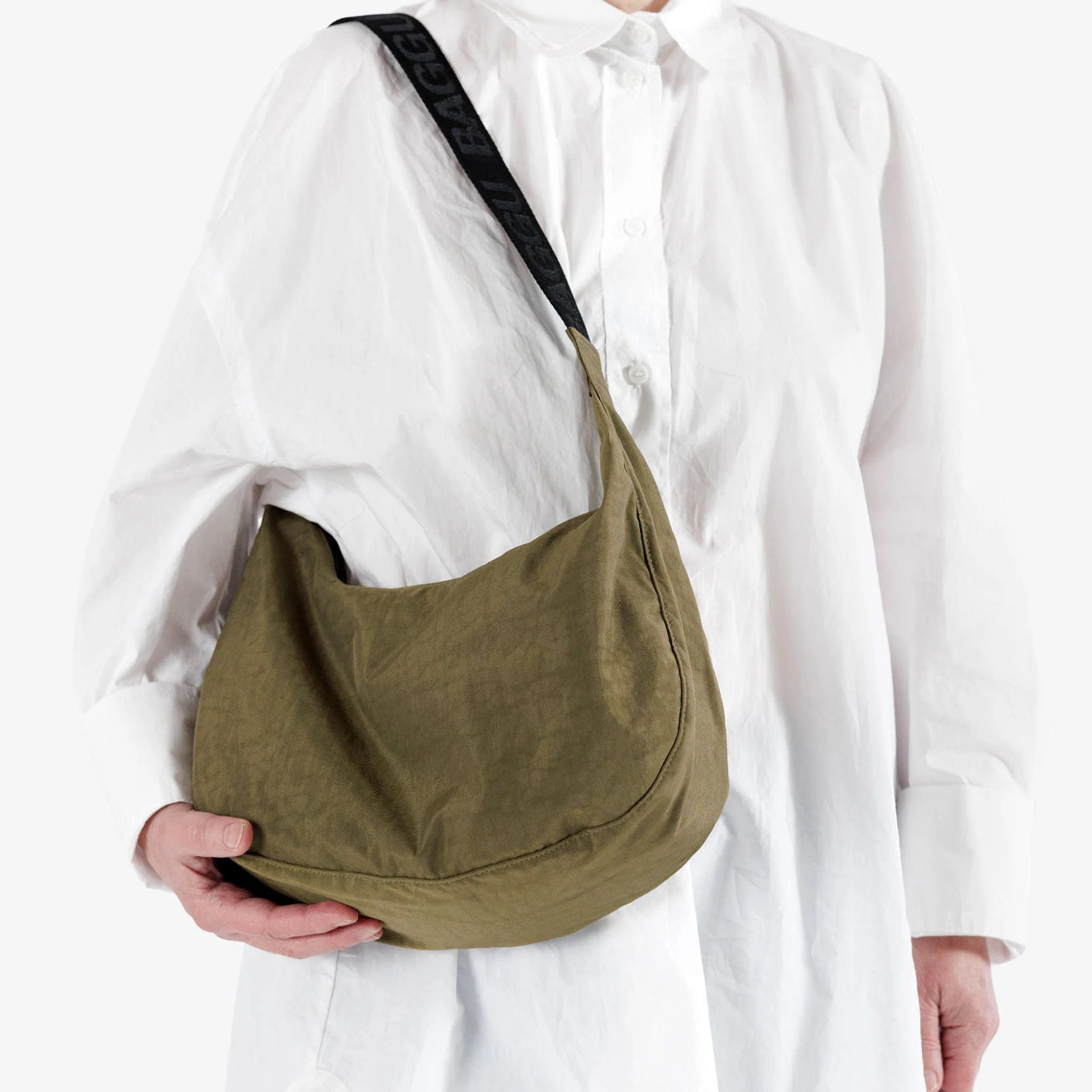 A person in a white shirt is holding the Baggu Medium Nylon Crescent Bag in Seaweed, featuring a sleek black strap that makes it perfect for crossbody wear.