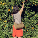 A person in a gray tank top and red shorts reaches for oranges, with the Baggu Medium Nylon Crescent Bag in seaweed green completing the ensemble.