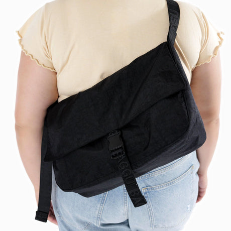 The person is wearing a light shirt and jeans, effortlessly carrying a spacious Baggu Messenger Bag - Recycled Nylon in black, perfectly designed to accommodate a laptop for any adventure.