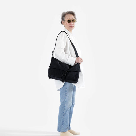 A person wearing sunglasses, a white shirt, light jeans, and holding a Baggu Messenger Bag - Recycled Nylon against a plain background.