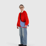 A person dressed in a red sweater, jeans, and sunglasses poses against a plain backdrop while holding the Baggu Medium Nylon Crescent Bag in Cornflower, crafted from recycled nylon.