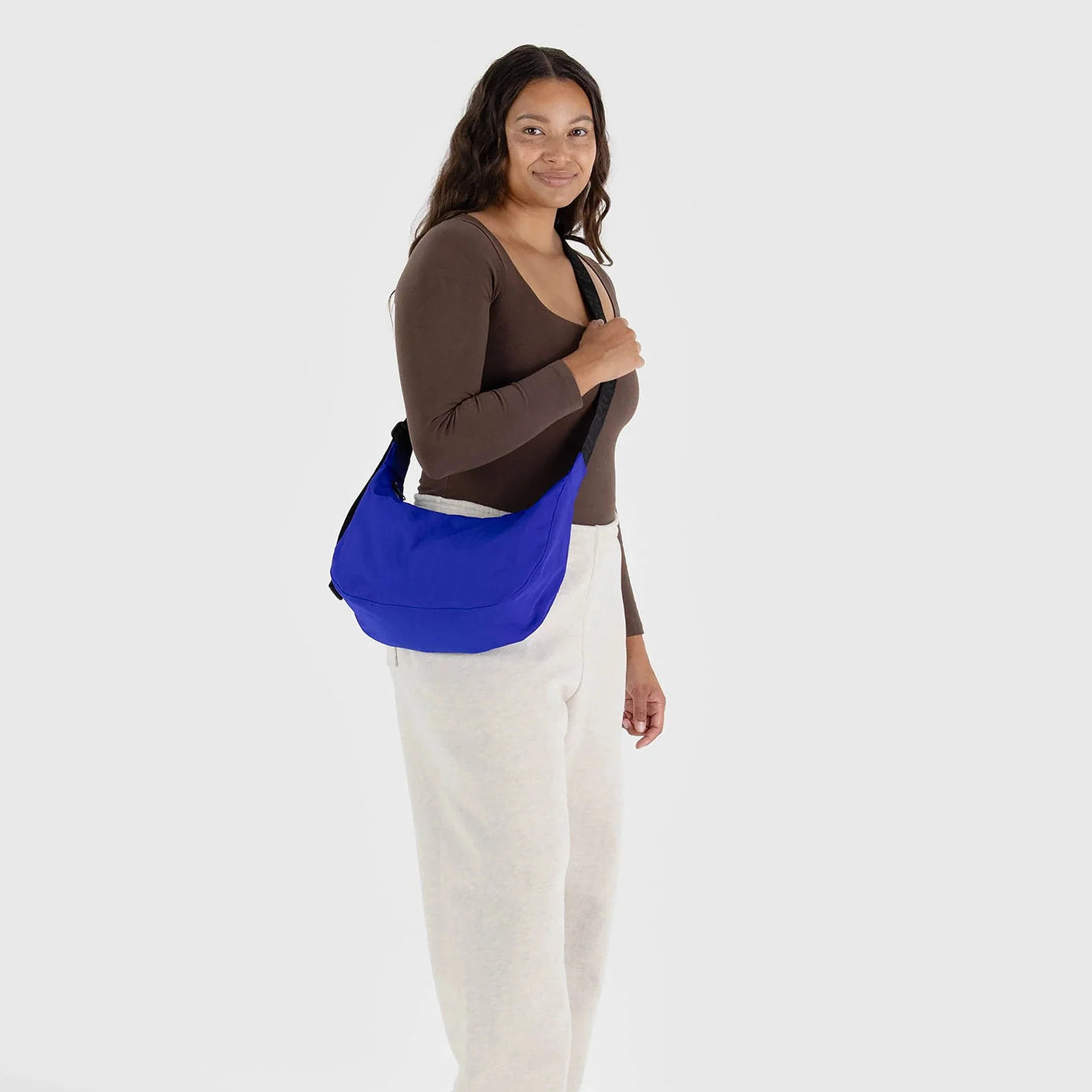 A person dressed in a brown top and white pants carries the Baggu Medium Nylon Crescent Bag in Lapis Blue, made from recycled nylon, set against a light gray background.