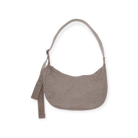 The Baggu Medium Nylon Crescent Bag in Dove is a beige shoulder bag made from recycled nylon, featuring an adjustable tonal logo strap and an attached fabric tag.