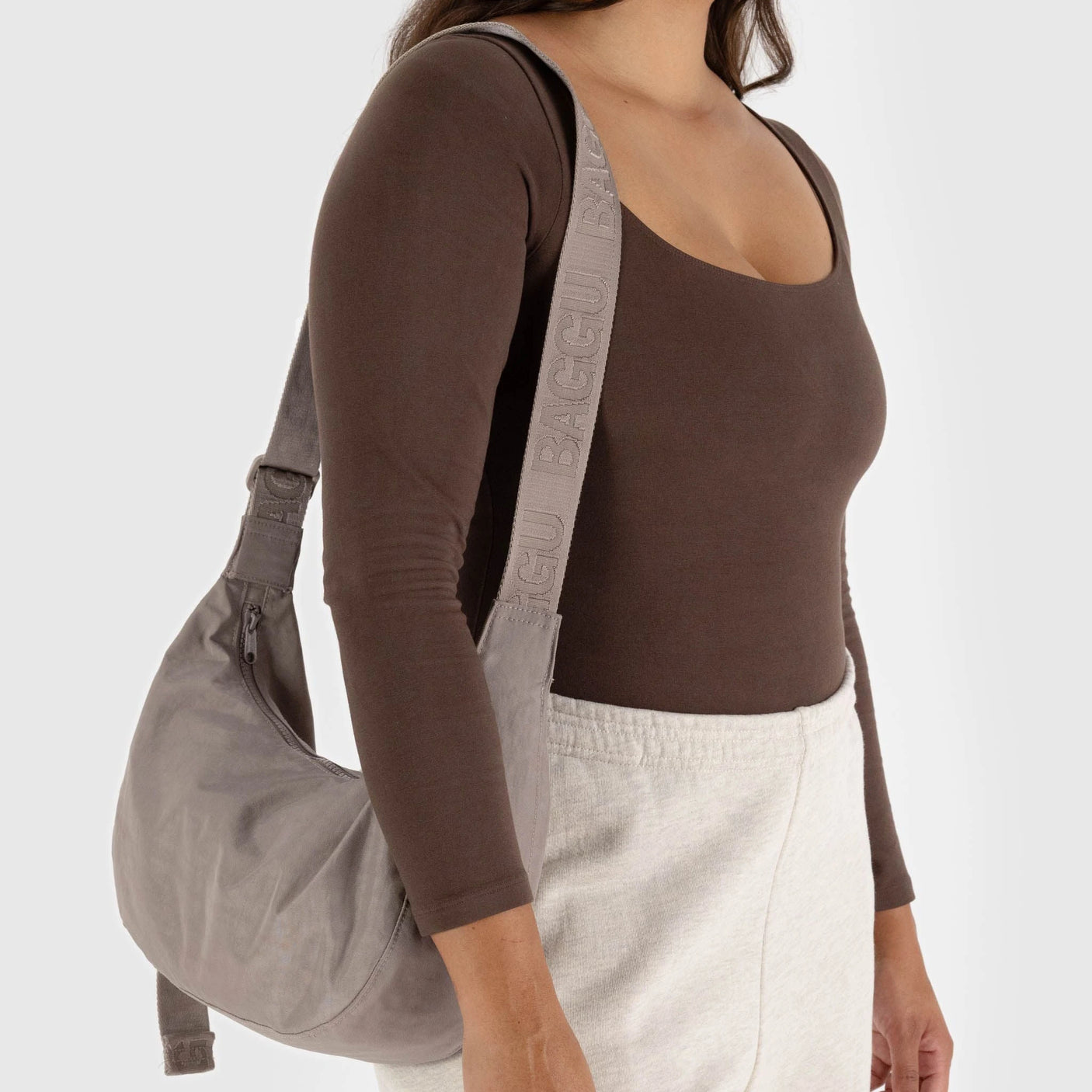 A person wearing a brown long-sleeve top and light-colored pants is carrying the Baggu Medium Nylon Crescent Bag in Dove, a sleek gray shoulder bag featuring an adjustable tonal logo strap. This stylish accessory is crafted from recycled nylon, adding an eco-friendly touch.