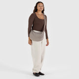 A person stands against a plain background, wearing a brown top, beige pants, and a Baggu Medium Nylon Crescent Bag in Dove with an adjustable tonal logo strap made from recycled nylon.