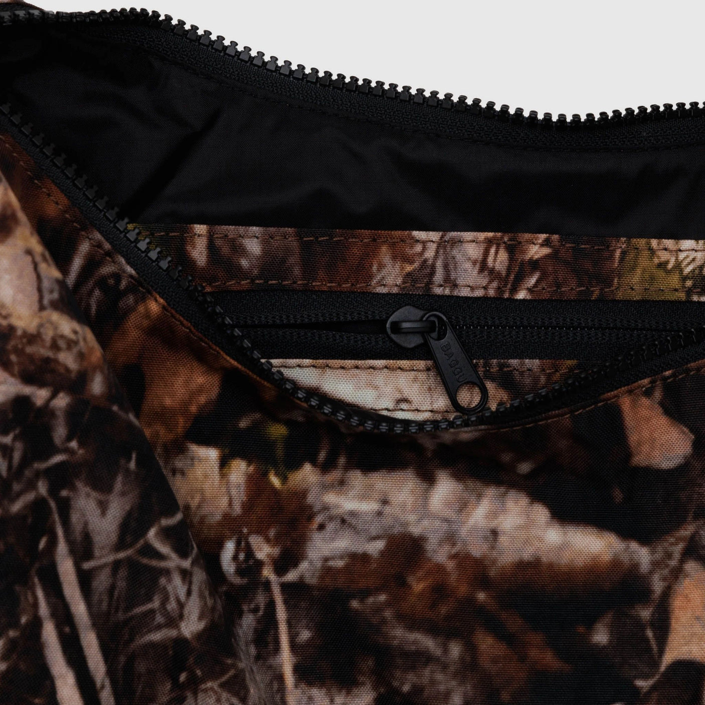 A detailed view of the Baggu Medium Nylon Crescent Bag in Photo Forest, featuring a camouflage-patterned fabric with a black zipper pocket, made from recycled nylon.