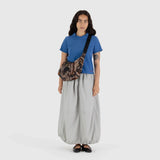 A woman stands with long hair, dressed in a blue polo shirt, gray skirt, and black shoes. She carries a camo-patterned Baggu Medium Nylon Crescent Bag in Photo Forest design, made from recycled nylon, all set against a plain white background.