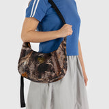 A person wearing a blue shirt and gray skirt is carrying the Baggu Medium Nylon Crescent Bag in the Photo Forest pattern.