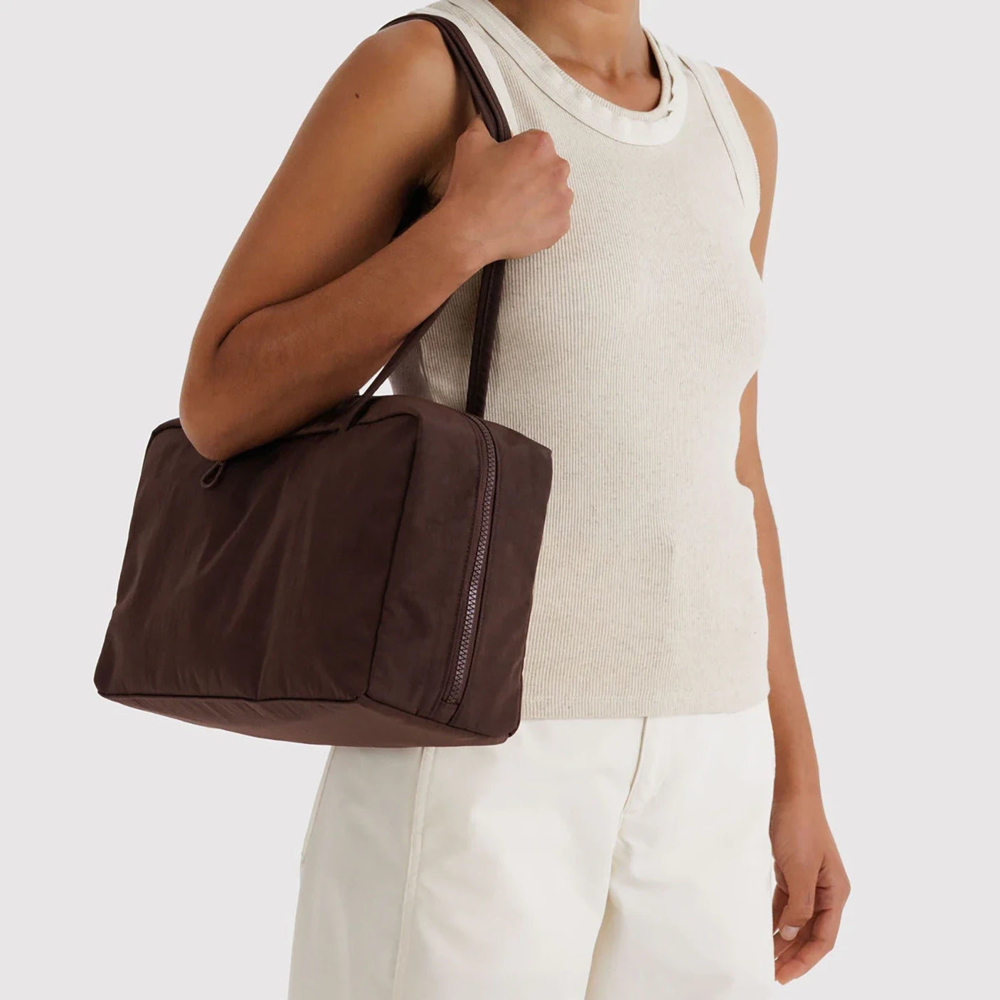 Dark brown rectangular fabric shoulder bag with dual straps and visible zipper held by person in sleeveless beige top and white pants
