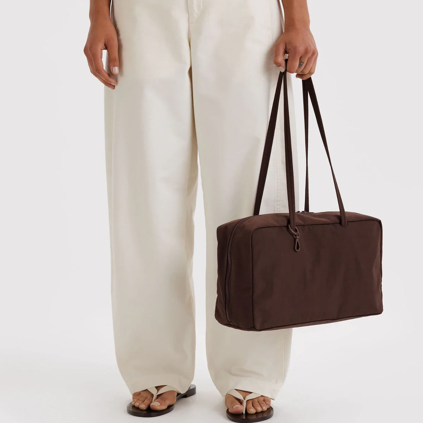 Dark brown rectangular fabric handbag with long dual straps and top zipper held by person in white wide-leg pants and sandals