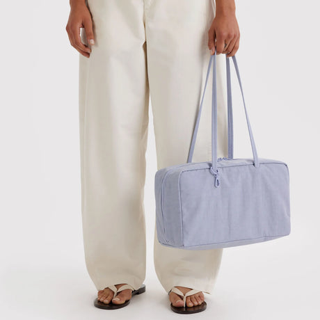 Lavender rectangular fabric tote bag with dual long straps and zipper closure held by person in white pants