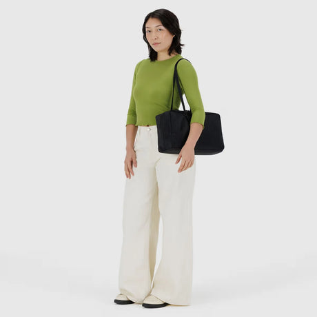 A person in a green long-sleeve shirt, white wide-leg pants, and black shoes stands holding the Baggu Medium Nylon Bowler Bag - Black by Baggu against a plain light background.