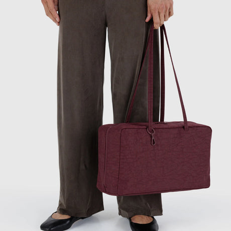 Person holding a mahogany medium nylon bowler bag by long straps against a light background