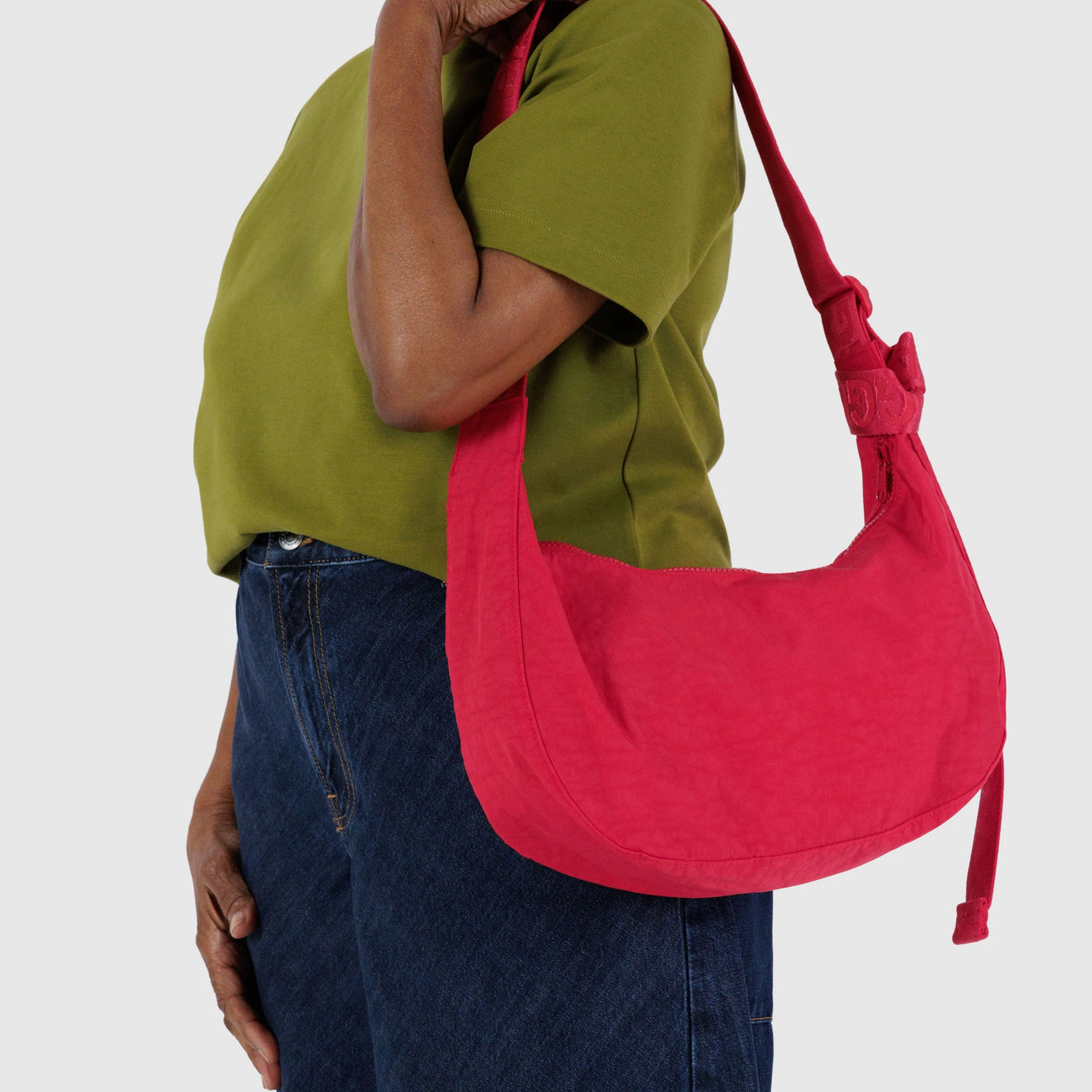 A person strolls by with a Baggu Medium Nylon Crescent Bag in Candy Apple, wearing a green shirt and blue jeans.