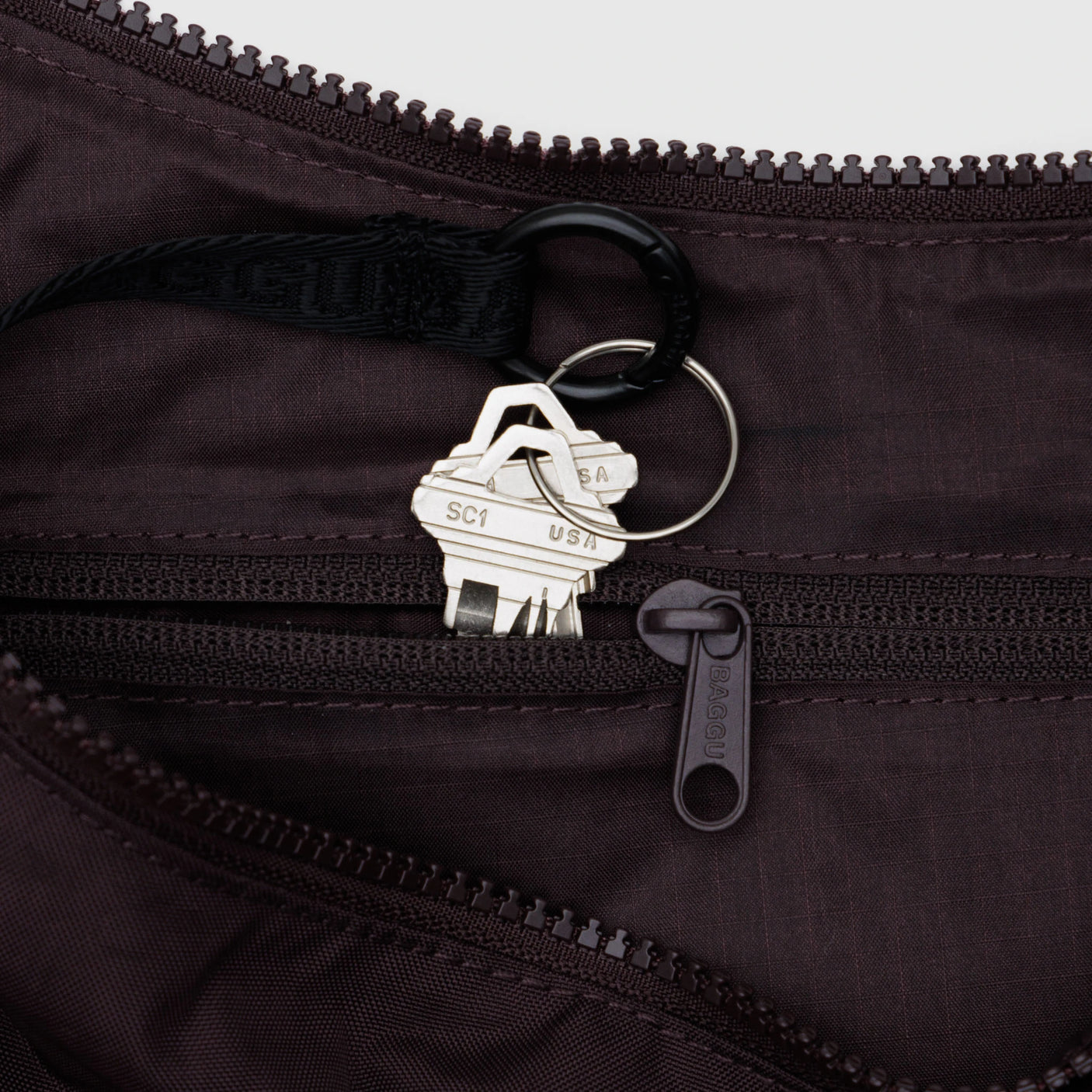 A set of keys on a black keyring is inside the brown zippered pocket of the Baggu Medium Nylon Crescent Bag in Chocolate Plum.