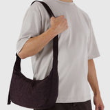 A person in a light gray T-shirt holds the strap of a Baggu Medium Nylon Crescent Bag - Chocolate Plum, highlighting its modern design and eco-friendly recycled nylon by Baggu.