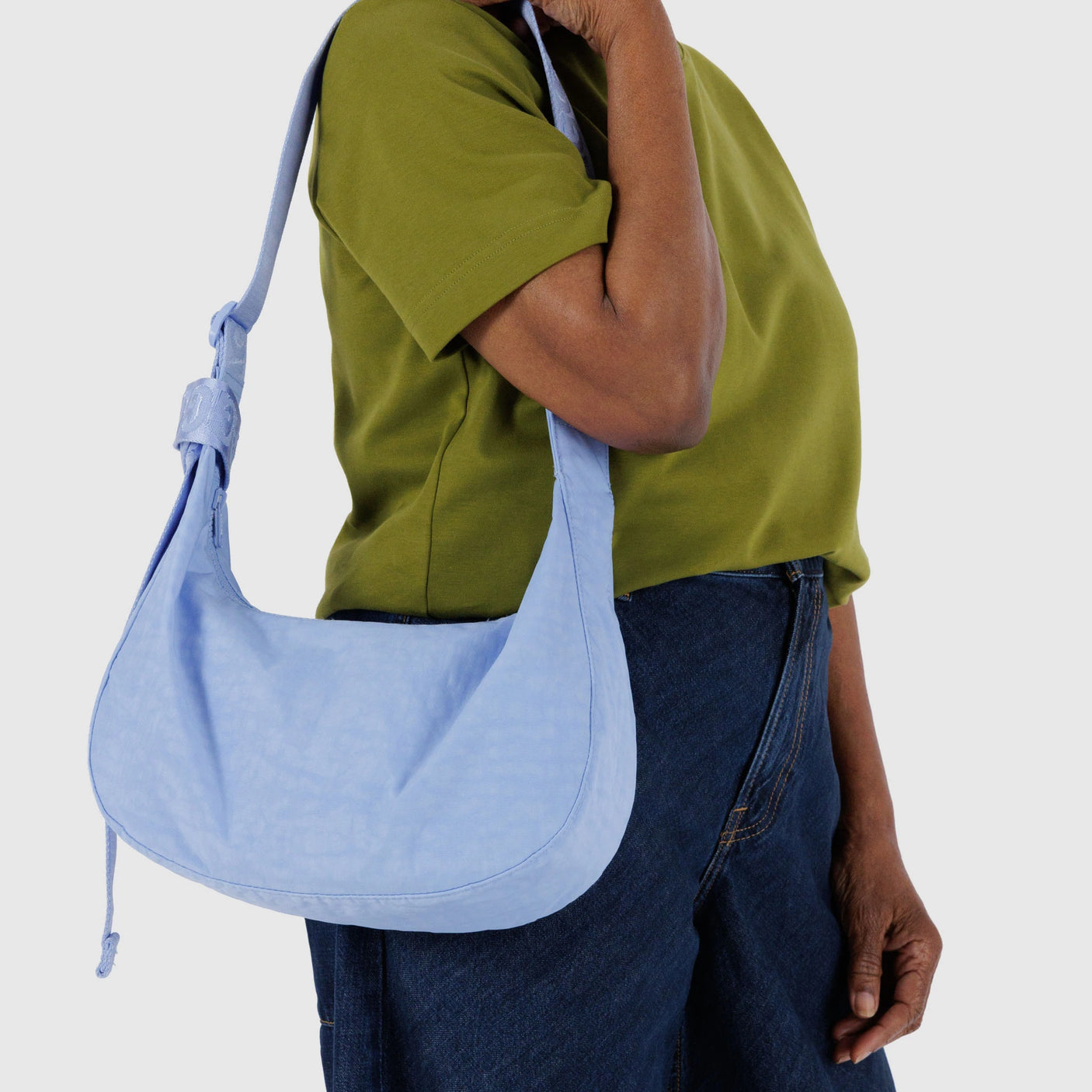 Against a plain background, a person in a green shirt and blue jeans holds the Baggu Medium Nylon Crescent Bag - French Blue, made from recycled nylon.