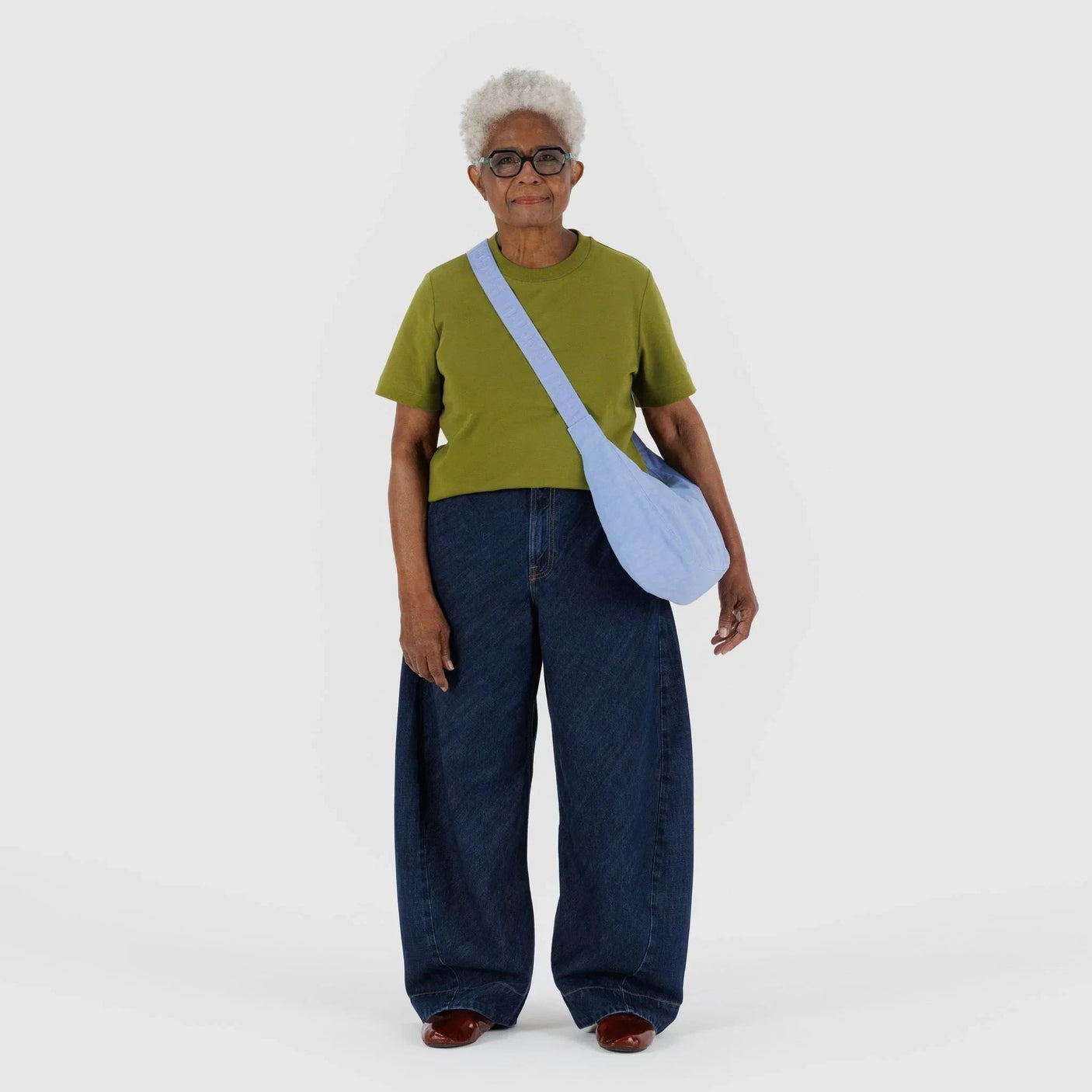 An older adult in a green shirt, wide-leg jeans, and brown shoes stands against a plain backdrop, carrying a Baggu Medium Nylon Crescent Bag in French Blue made of recycled nylon.