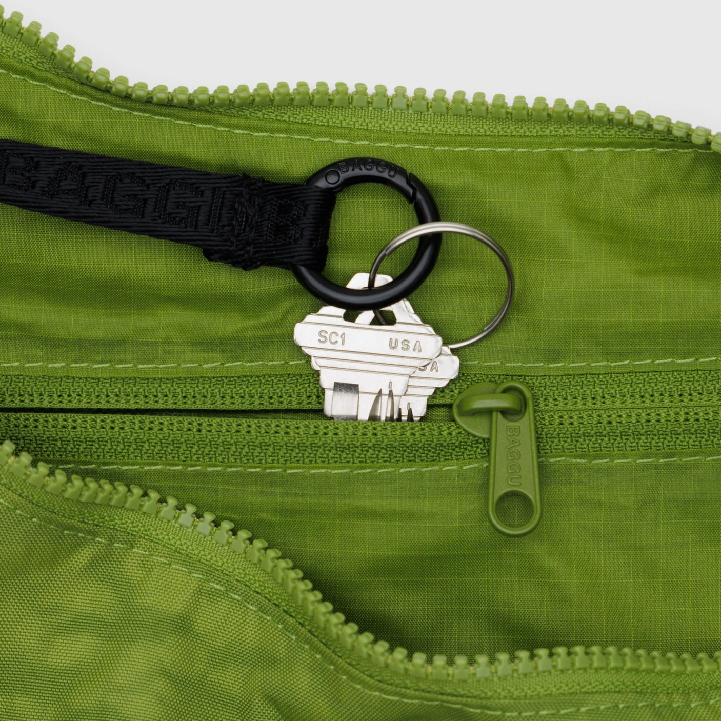 A set of silver keys dangles on a black ring inside a vibrant green zippered pouch, reminiscent of the sleek compartments in the Baggu Medium Nylon Crescent Bag - Green Juice. Two zippers are visible, subtly suggesting an eco-friendly lifestyle by Baggu.