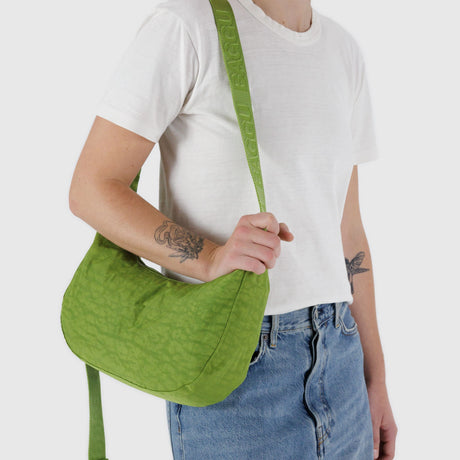 A person in a white T-shirt and blue jeans carries the Baggu Medium Nylon Crescent Bag in Green Juice, sipping on a refreshing green juice.