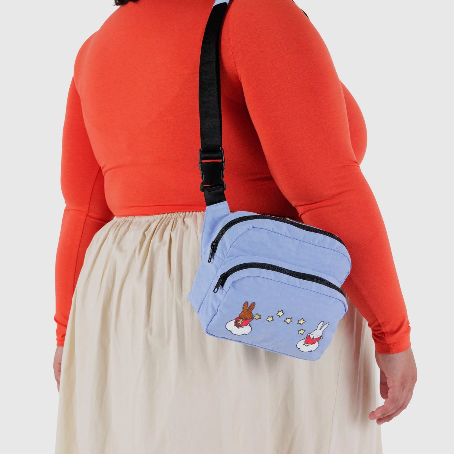 A person in an orange long-sleeve top and beige skirt wears a light blue Baggu Miffy x Baggu Fanny Pack featuring cartoon bunnies and stars, made from recycled nylon.